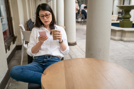 relaxed asian Taiwanese female wearing earphones watching online clip videos on mobile phone while having coffee break at a cafe in palo alto city California usaの写真素材