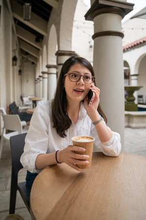 smiling asian Japanese woman having phone discussion with business partner at the outdoor seating of downtown coffee shop in palo alto city California usaの写真素材
