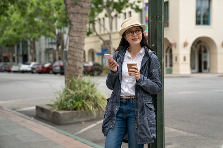 happy asian Taiwanese woman tourist carrying coffee and enjoying beautiful cityscape in downtown palo alto while using cellphone for online guide on the corner of street in California usaの写真素材