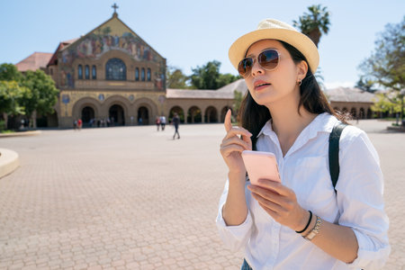 confused asian korean girl traveler using online map on phone and looking into space with finger gesture near Stanford Memorial Church, not sure where another landmark is at Stanford universityの写真素材