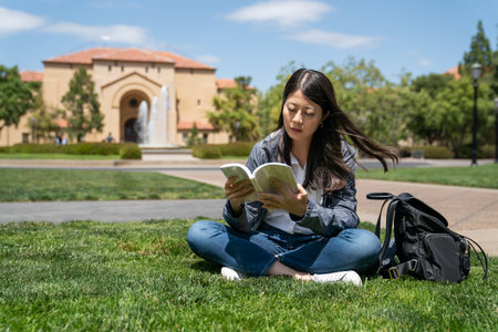 concentrated asian Taiwanese girl international student sitting on grass and studying textbook on a sunny day on Stanford campus with fountain at backgroundの写真素材