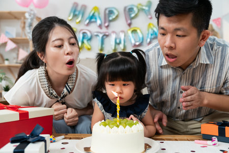 closeup of cute asian baby girl making birthday wishes with father and mother before blowing out candle on her second birthday party at homeの写真素材