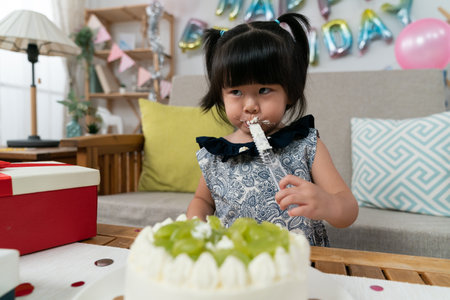 selective focus of cute naughty asian baby girl licking yummy cream on cake server during her second birthday celebration at homeの写真素材