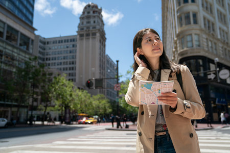 pretty asian korean female traveler carrying route map and tucking hair on Market Street while enjoying sunny weather and cityscape in san Francisco California usaの写真素材