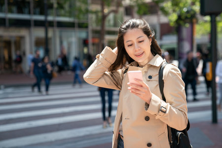 cheerful asian Taiwanese girl tourist pushing hair back and reading online travel guide on cellphone on street corner while visiting san Francisco in California usaの写真素材