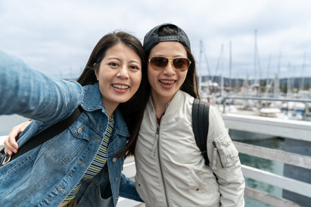 two smiling asian girls looking at camera taking selfie picture while on vacation at Old Fisherman Wharf in California. one wearing sunglasses putting hand around her friendの写真素材