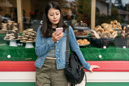 asian Taiwanese female looking at her cellphone while waiting for friend outside a neighborhood pastry shop in Carmel by the sea against display window backgroundの写真素材