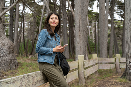 smiling asian chinese woman traveler sitting relaxing on low fence and enjoying natural beauty while visiting the crocker grove along 17 mile drive in California usaの写真素材