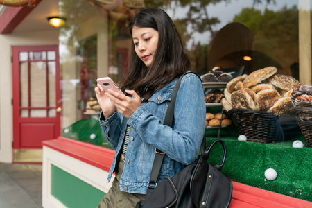 leisure asian Japanese girl traveler searching travel guide on the phone while taking a break outside a bread bakery during her stay at Carmel by the sea in California usaの写真素材