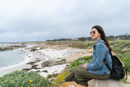 leisure asian chinese girl traveler wearing sunglasses sitting at beachside to enjoy beautiful ocean view on a breezy day during road trip along state route one in California usaの写真素材