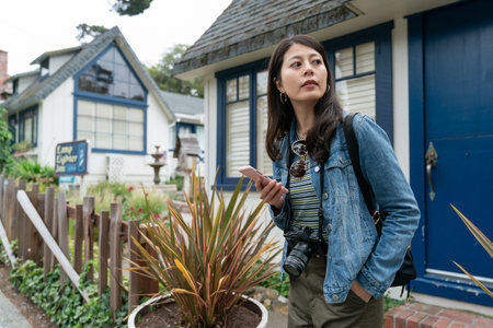 asian Chinese girl visitor using phone to check time and looking away into distance while waiting for friend near a white cottage house with blue door in Carmel by the seaの写真素材