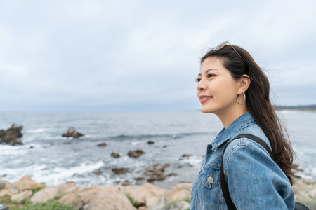 closeup portrait of pretty smiling asian girl looking into space at scenic ocean view at an lookout area in the wind during her road trip on California state route one in usaの写真素材