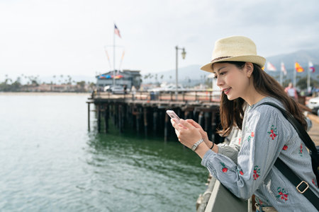 portrait of pretty asian Japanese girl wearing hat and enjoying social media on phone while relaxing on waterfront railing at santa Barbara harbor in california usaの写真素材