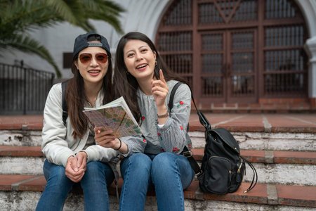 two smiling asian chinese women visitors looking at beautiful government buildings with pointing gesture while consulting map on stone stairs at santa Barbara courthouseの写真素材