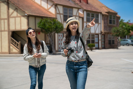 cheerful asian chinese female visitor carrying camera and pointing at famous landmark in space as her friend is looking with curiosity behind near Danish country style homes in solvangの写真素材