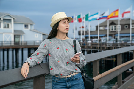 portrait of asian taiwanese female traveler resting arm on pier railing and gazing into distance while using phone for travel info at santa Barbara harbor in california usaの写真素材