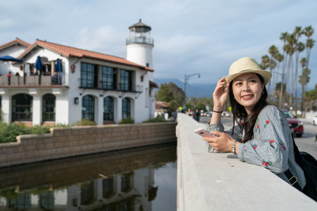 leisure asian Chinese female traveler holding hat leaning on stone railing and looking into distance at nice view in santa Barbara with a lighthouse at background at duskの写真素材
