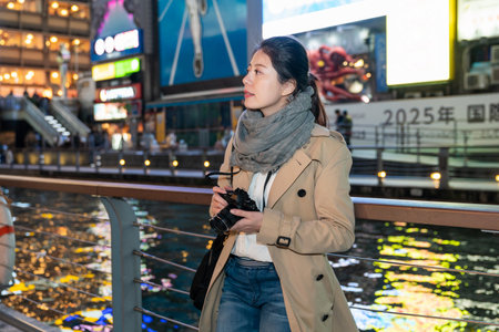 attractive Asian Chinese female photographer looking into space feeling the vibe of Shinsaibashisuji shopping area at night while leaning against handrail by doutonbori river in Osaka japanの写真素材