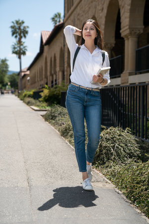 full length of smiling asian chinese girl exchange student carrying textbook and pushing hair back while enjoying sun and breeze on her way to class at school university in California usaの写真素材