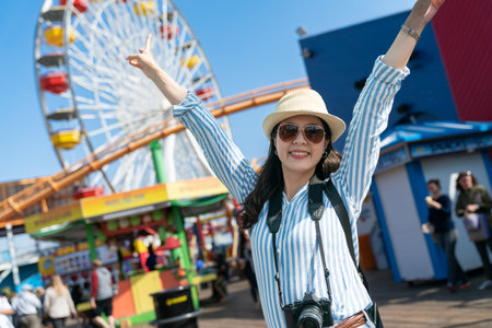 happy stylish asian Japanese female visitor on holiday at amusement park looking at camera with raised arms and victory hand signs on background of a Ferris Wheelの写真素材