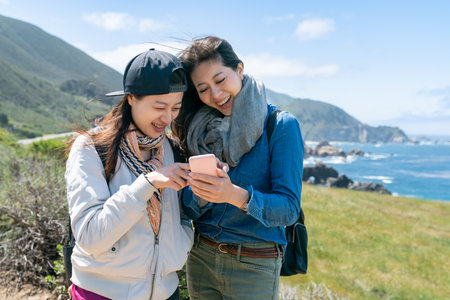 two cheerful asian Korean sister tourists looking at guide on phone together and discussing famous...