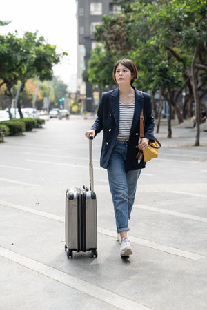 full length portrait of asian Korean businesswoman pulling baggage and looking around surroundings while traveling for work in city. solo business trip themeの写真素材