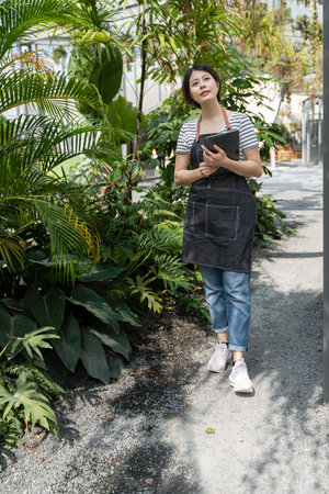 asian Korean young female gardener looking up enjoying sunny weather while working in tropical glasshouse with an electronic padの写真素材