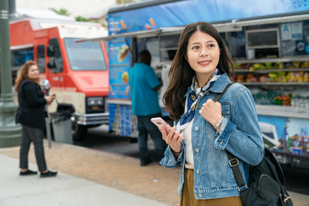 Asian female engineer holding mobile phone looking away with smile in front of food truck during lunch break in USAの写真素材