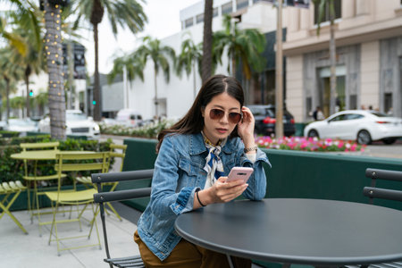 young asain woman sitting at a table and typing text messages on smartphone at a bar or restaurant outdoorsの写真素材
