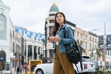 stylish Asian Japanese businesswoman carrying phone and looking into distance while crossing the busy street in downtown Los angels California usa with cars at backgroundの写真素材
