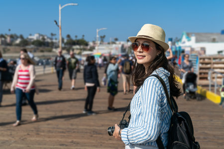 beautiful smiling asian korean girl wearing sunglasses and backpack turning to look at distance at santa monica pier with tourists coming and going at backgroundの写真素材