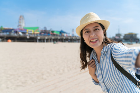 self portrait of happy asian Japanese girl tourist wearing hat looking at camera with smile on background of santa monica beach against blue skyの写真素材
