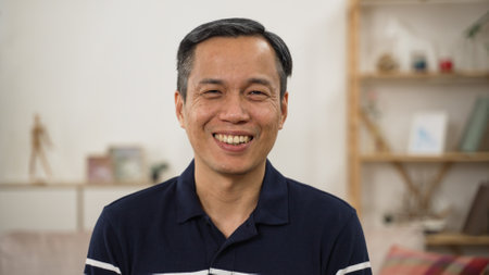 shoulder shot portrait of happy asian senior man looking at the camera with a big smile against modern cozy home backgroundの写真素材