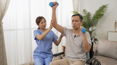 asian paralyzed elderly man sitting in wheelchair and going through physical therapy with personal care attendantâs support. he does weight lifting using dumbbells at homeの写真素材