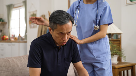 closeup of asian senior man is coughing while home attendant is patting on his back to help loose the sputum from lung in living room at home. domiciliary care conceptの写真素材
