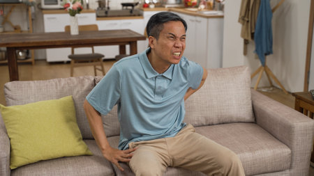 Chinese mature man moving to sit and rest on sofa while suffering pain in lower back and moaning with great discomfort. Elder senior person slip and fall risk at home conceptの写真素材