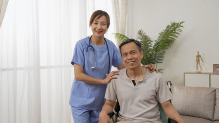 portrait of smiling asian resident care attendant and disabled mature man on wheelchair posing and looking at camera near the window at home with daylightの写真素材
