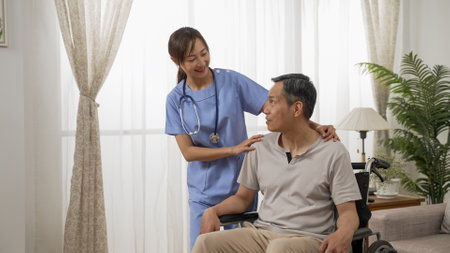 cheerful asian resident care attendant smiling while chatting with older man on wheelchair and encouraging him near the window at home during the dayの写真素材