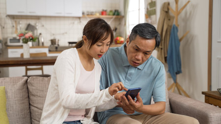 confused asian grandpa asking adult granddaughter questions while using cellphone in living room. the woman puts down her phone and teaches him how to useの写真素材