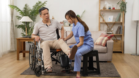 asian female care attendant helping senior man on wheelchair doing rehab training. she slowly lifts his injured leg while he shows painful face in the living room at homeの写真素材