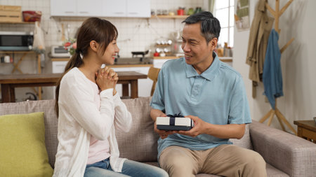 smiling asian daughter giving happy father a surprise present for Fatherâs Day celebration at home. she removes hand from his eyes and hopes he likes the giftの写真素材