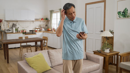 Forgetful Japanese mature man finding his smartphone behind sofa cushion at home. He scratches his head and talks to himself feeling embarrassed and sits down to use itの写真素材