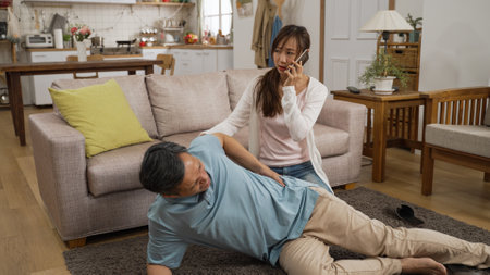 Asian senior father suffering pain from a fall and lying on living room floor with a stick beside at home. Her adult daughter quickly comes over to help and calls 911 on cellphoneの写真素材