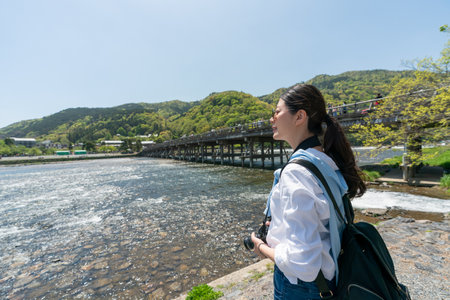 asian Japanese girl backpacker looking at calm Katsura river under sunny sky and enjoying tranquility with Togetsu-kyo Bridge at background while visiting arashiyama in Kyoto japanの写真素材