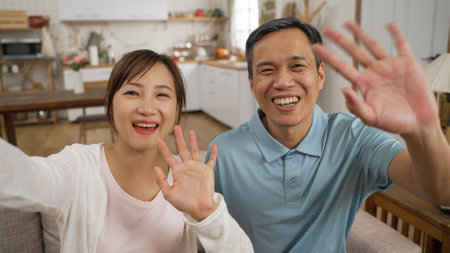 mobile cam view of cheerful asian Taiwanese adult daughter and father waving hi to camera and greeting their friends with smile while having online video chat at homeの写真素材