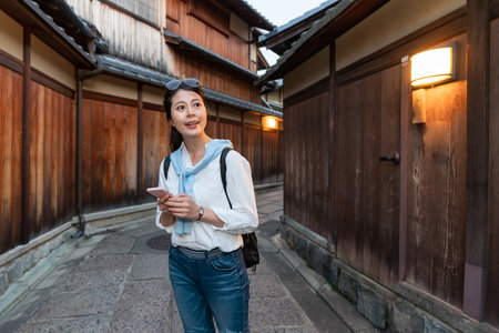 cheerful asian taiwanese female tourist learning travel information on phone and admiring ancient wooden architectures lining up Ishibeikoji lane in gion Kyoto japan at duskの写真素材
