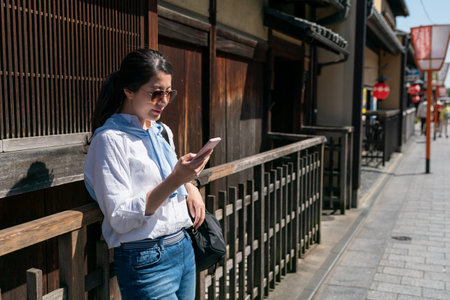fashionable asian Korean girl tourist looking at her cellphone while leaning relaxing at the exterior of wooden Japanese teahouse on Gion Hanamikoji Street in Kyoto japan on sunny dayの写真素材