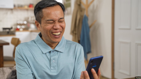 cheerful asian mature male enjoying watching funny online video on his smartphone at home. he points at phone screen while laughingの写真素材