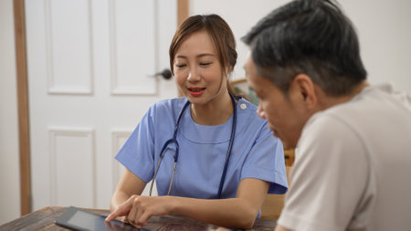 smiling Asian woman nursing aide showing treatment plan to senior male patient using digital pad during home visit. she explains with hand gesturesの写真素材