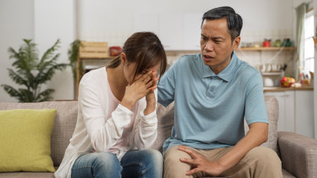 asian Japanese grandfather trying to comfort and encourage sad adult granddaughter on sofa at home. he taps her back while she covers her face feeling frustrationの写真素材